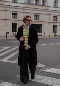 Long manteau noir sur un pull rayé vert et blanc, pantalons gris plissés et chaussures noires. Des lunettes et des accessoires complètent le look.