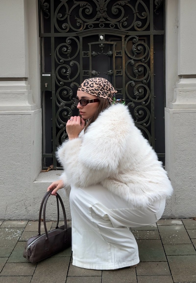 Donna con cappotto di pelliccia bianca, pantaloni bianchi, foulard leopardato e occhiali da sole, accovacciata sul marciapiede mentre tiene una borsa marrone accanto a un cancello nero ornato.