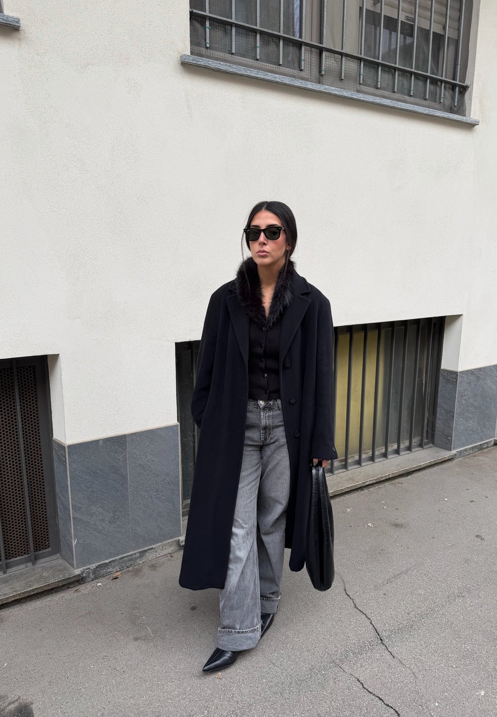 Manteau en laine noire avec col en fourrure, haut noir boutonné, jean gris à jambes larges, et chaussures noires à bouts pointus. Accessoires : un sac à main noir.