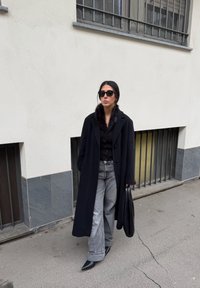Manteau en laine noire avec col en fourrure, haut noir boutonné, jean gris à jambes larges, et chaussures noires à bouts pointus. Accessoires : un sac à main noir.