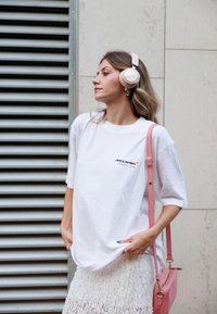 Λευκό oversized μπλουζάκι με το λογότυπο "McLaren", σε συνδυασμό με μια υφασμάτινη λευκή φούστα. Φοράει ροζ ακουστικά που καλύπτουν τα αυτιά και κρατάει μια ροζ τσάντα.