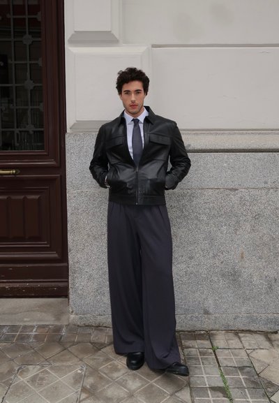 Chaqueta de cuero negro con cremallera y bolsillos con solapa, combinada con pantalones grises de gran tamaño y zapatos negros. Hombre de pie contra una pared texturizada.