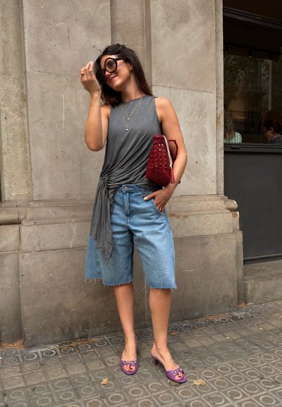 Mujer que lleva gafas de sol grandes, una camiseta gris sin mangas, pantalones cortos vaqueros, tacones morados y sostiene una bolsa roja, de pie contra una pared de piedra.
