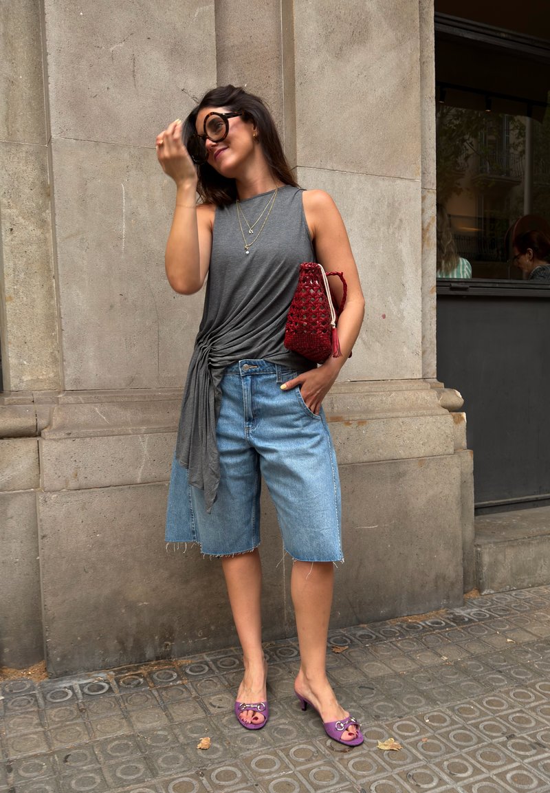 Femme portant des lunettes de soleil surdimensionnées, un haut gris sans manches, un short en jean, des talons violets et tenant un sac rouge, debout contre un mur en pierre.