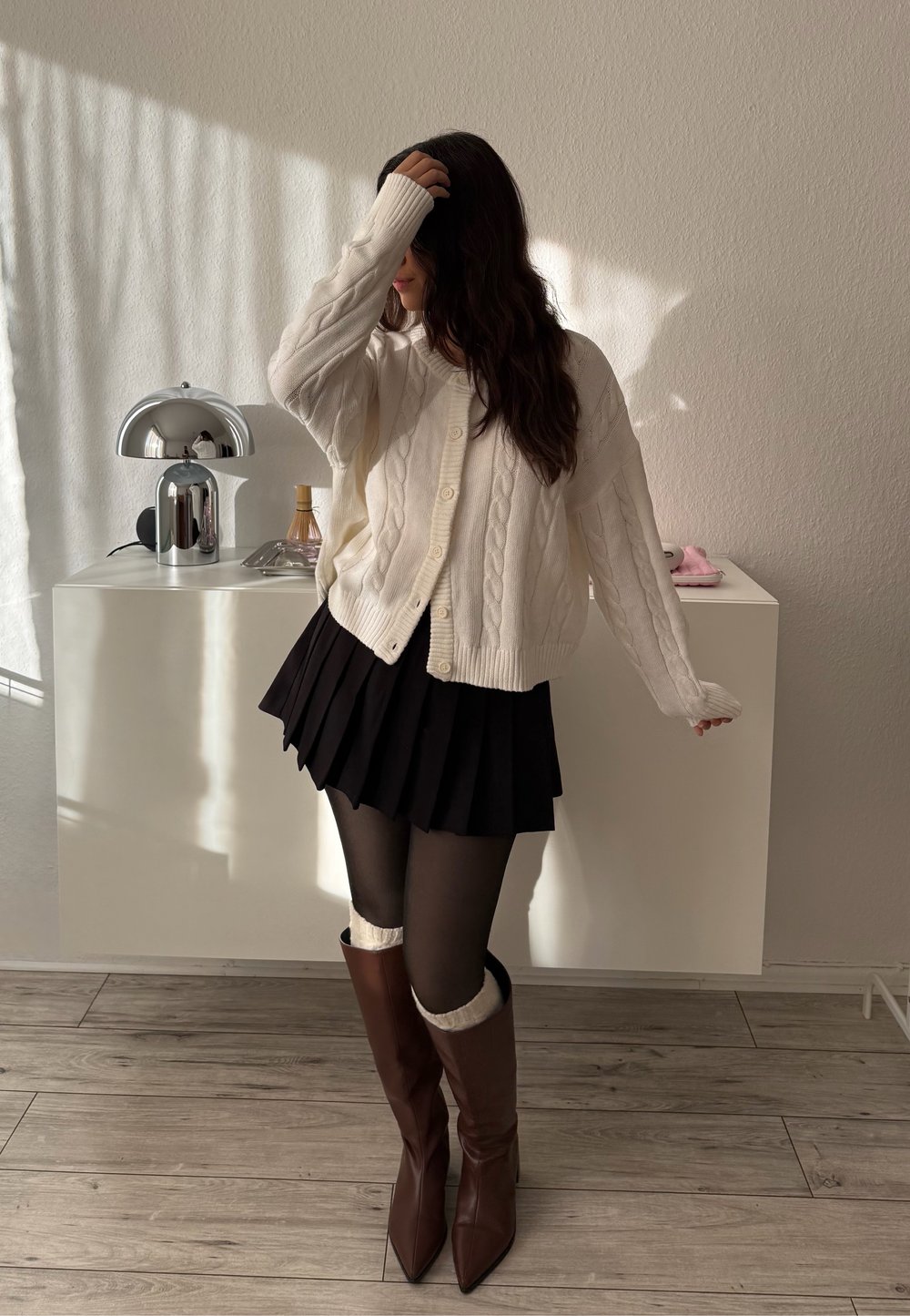 Femme aux longs cheveux bruns portant un cardigan en maille torsadée blanc, une jupe plissée noire, des collants noirs et de longues bottes brunes, debout près d'un meuble blanc.