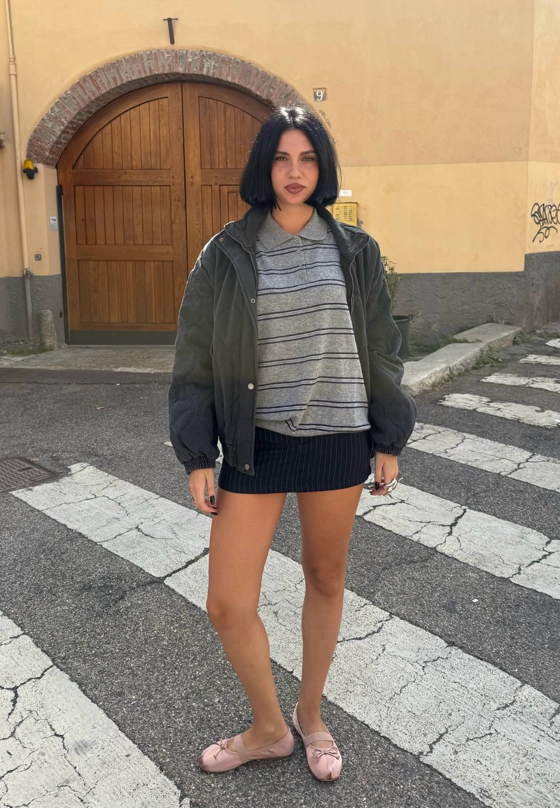 Giovane donna con capelli neri corti, che indossa una polo a righe, una giacca scura, una gonna nera a righe sottili e ballerine rosa, in piedi su un attraversamento pedonale.