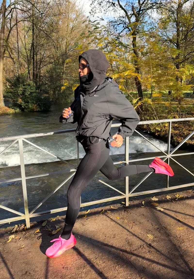 Veste noire à fermeture éclair avec capuche, leggings noirs texturés et chaussures de course roses vives. Joggeur en mouvement le long d'un chemin près de l'eau.