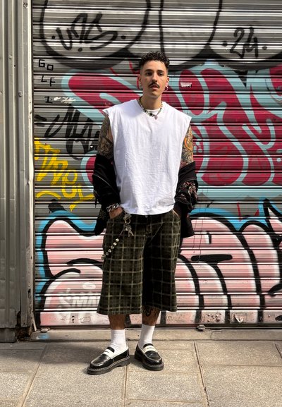 Camiseta sin mangas blanca, chaqueta oversized negra, pantalones cortos de cuadros verdes, zapatos negros con calcetines blancos, joyería plateada y tatuajes en los brazos.