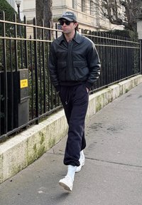 Άνδρας που φοράει μαύρο δερμάτινο μπουφάν, σκούρα φόρμα, λευκά αθλητικά παπούτσια, καπέλο και γυαλιά ηλίου περπατάει σε πεζοδρόμιο της πόλης δίπλα σε μεταλλικό φράχτη.