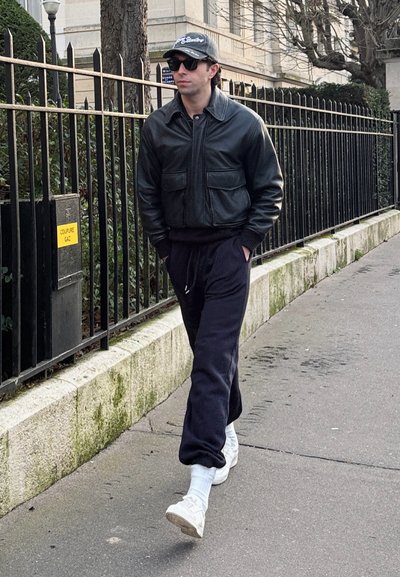 Hombre vestido con chaqueta de cuero negra, pantalones deportivos oscuros, zapatillas blancas, gorra y gafas de sol caminando por una acera de la ciudad junto a una valla metálica.