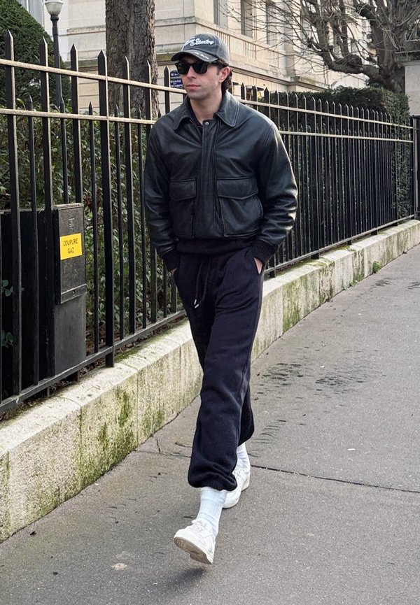 Férfi fekete bőrdzsekiben, sötét szabadidőnadrágban, fehér tornacipőben, sapkában és napszemüvegben, amint egy városi járdán sétál egy fémkerítés mellett.