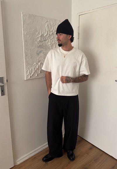 Camiseta blanca de manga corta, pantalones negros holgados, gorro negro y zapatos negros, con una obra de arte mural abstracta texturizada de fondo.