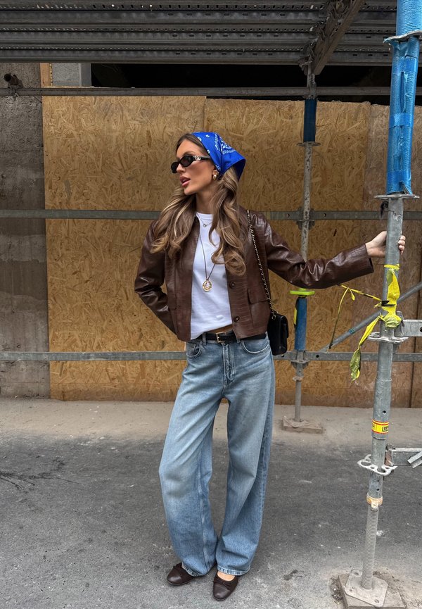 Mujer con chaqueta de cuero marrón, camisa blanca, vaqueros anchos azules, pañuelo azul en la cabeza y gafas de sol, de pie sosteniendo un poste de andamio de construcción.
