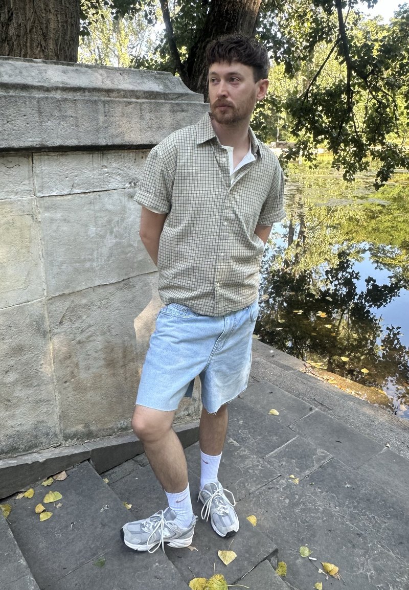 Jeune homme en chemise à manches courtes à carreaux, short en denim et baskets, debout sur des marches en pierre près d’un étang avec des arbres et des reflets.
