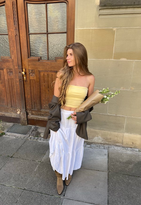 Giovane donna con capelli lunghi in piedi che tiene fiori avvolti, indossa un top giallo, una gonna bianca, stivali marroni e una giacca marrone sulle braccia.