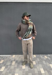 Sweat à capuche marron, écharpe à carreaux verts avec des rayures orange, pantalon en velours côtelé beige, sabots olives, lunettes de soleil foncées, fond mur gris.