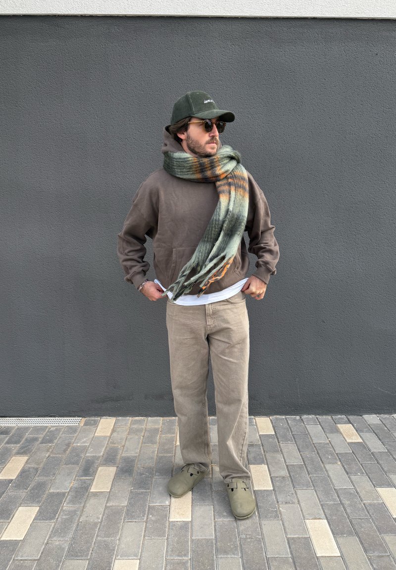 Brown hoody, green checked scarf with orange stripes, beige corduroy trousers, olive clogs, dark sunglasses, grey wall background.