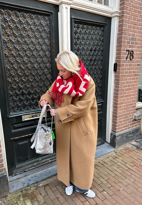 Manteau beige oversize, écharpe rouge avec motif graphique, sac argenté, jeans bleu clair, chaussures blanches à enfiler, debout devant une porte noire.