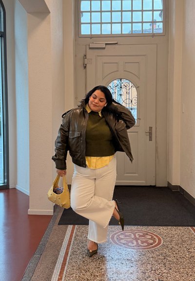 Mujer de pie en el interior cerca de una puerta, vestida con una chaqueta de cuero oscura, suéter verde, camisa amarilla, pantalones blancos y sosteniendo un bolso amarillo.