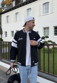 Schwarze und weiße Collegejacke mit gesticktem "Starter"-Logo, kombiniert mit einem weißen T-Shirt und hellblauen Jeans. Hundeleine in der Hand.