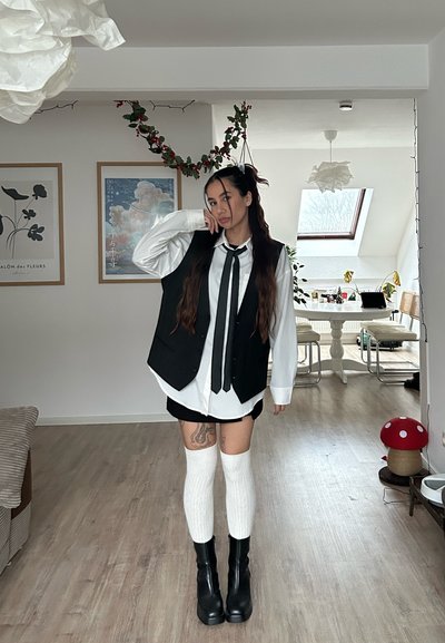 Chaleco negro sobre camisa blanca con corbata, falda negra, calcetas blancas hasta la rodilla y botas negras con plataforma. Ambiente interior con vegetación.