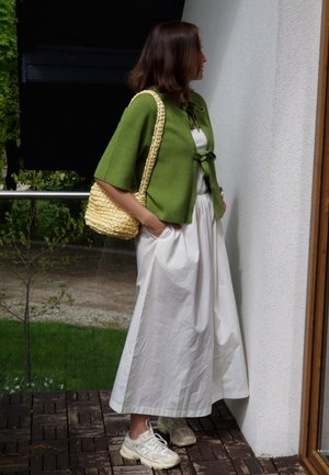 Green knit cardigan with short sleeves, tied at the front; white long skirt; woven straw bag; light sneakers. Natural setting background.