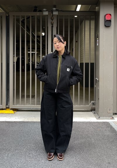 Chaqueta negra con cremallera sobre un suéter verde oliva, combinada con pantalones anchos negros y zapatos marrones. Tejido liso, ajuste relajado, detalles mínimos.