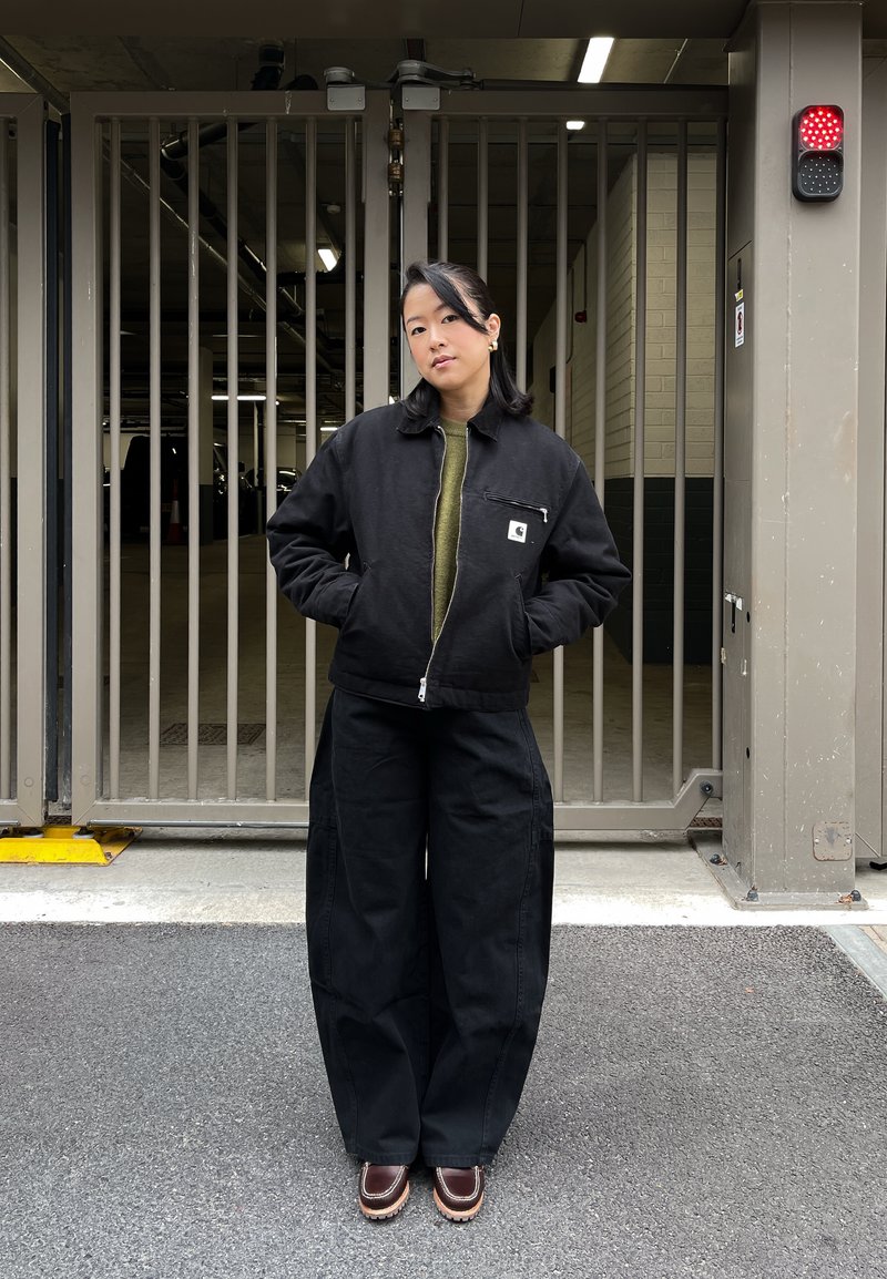 Veste noire zippée portée sur un pull vert olive, associée à un pantalon large noir et des chaussures marron. Tissu lisse, coupe décontractée, détails minimaux.