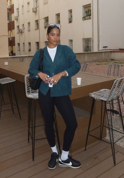 Cárdigan de felpa color verde azulado, parte superior blanca, leggings negros y zapatillas negras. Sosteniendo una taza, de pie junto a una mesa de madera con taburetes de metal.