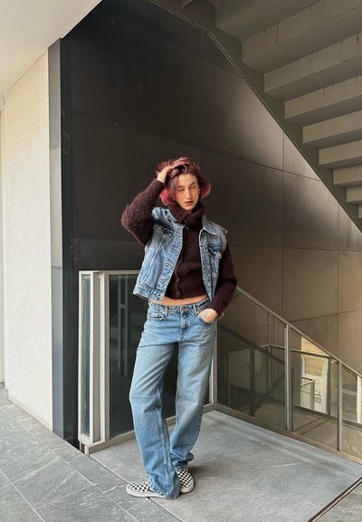 Chaleco de denim sobre un suéter de punto oscuro, combinado con jeans de color azul claro y zapatillas de cuadros en blanco y negro. De pie contra una escalera moderna.