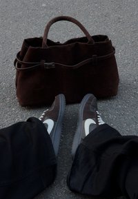 Geantă maro din piele întoarsă cu un design structurat și mâner superior, asortată cu pantaloni negri și adidași gri-maro cu un swoosh alb.