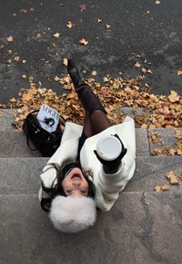 Cappello di pelliccia bianco, guanti neri, cappotto color crema e stivali neri alti fino al ginocchio. Tiene in mano una tazza di caffè, con una rivista Vogue e foglie autunnali vicino.