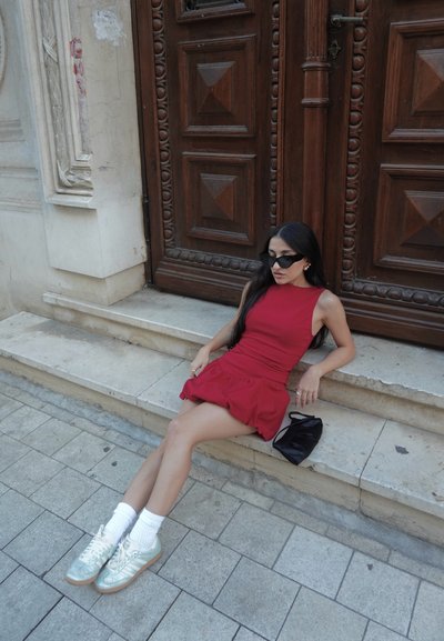 Vestido rojo sin mangas con un dobladillo con volantes, combinado con calcetines blancos y zapatillas de deporte brillantes en color menta. Un bolso negro descansa en los escalones de piedra cerca de una puerta de madera.