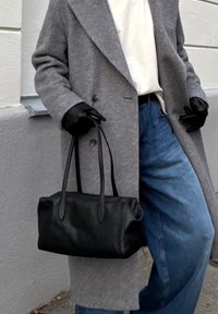 Mala de mão em couro preto com formato arredondado e duas alças, combinada com um casaco cinza de padrão espinha de peixe e jeans azuis de perna larga. Luvas pretas acentuam o visual.