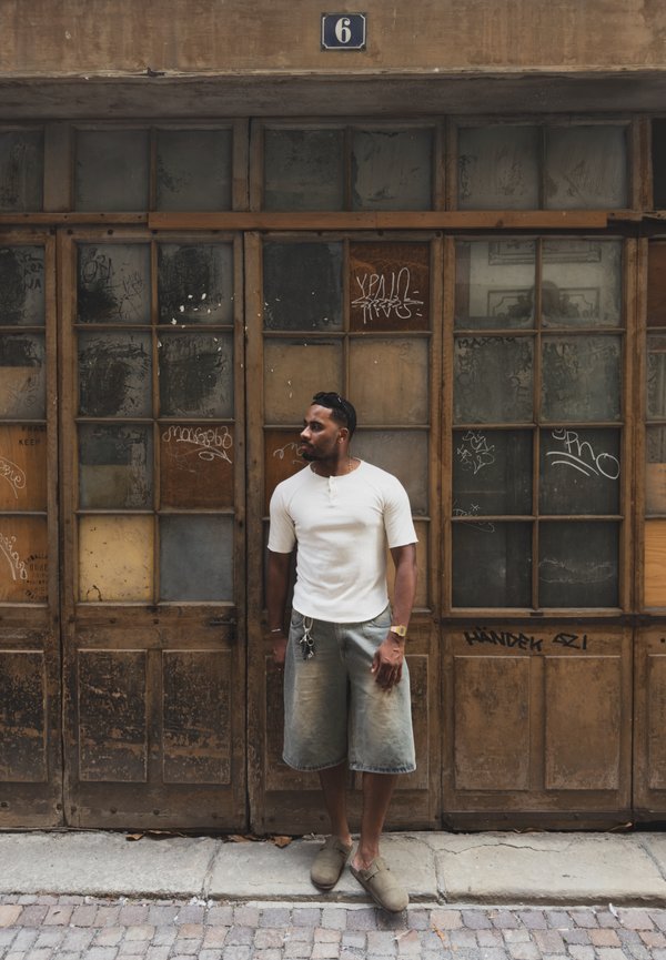 Homme en chemise blanche et bermuda long debout contre une vieille porte en bois avec des panneaux en verre et des graffitis, panneau numéro six au-dessus.