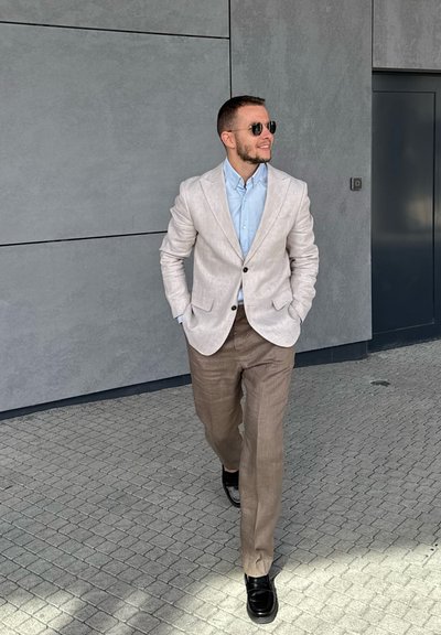 Blazer de lino gris claro sobre una camisa azul clara y pantalones color beige. Zapatos negros. Textura suave con un mínimo de detalles. Fondo de una pared gris.