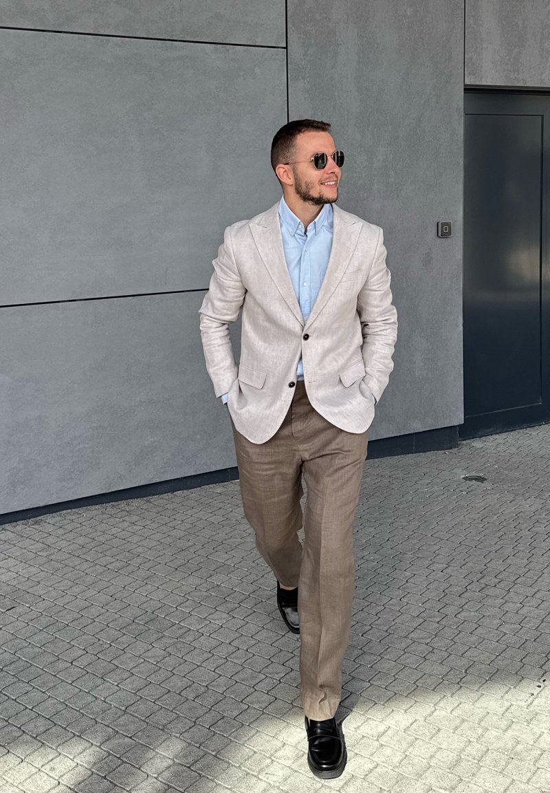 Šviesiai pilkas lino švarkas ant šviesiai mėlynos marškinių ir smėlio spalvos kelnių. Juodi batai. Lygus tekstūra su minimaliais akcentais. Fone - pilka siena.