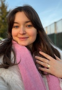 Giovane donna con lunghi capelli castani che indossa una sciarpa rosa sopra una giacca chiara, tiene la mano sul petto mostrando un anello con dettagli.