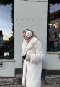 Bianco cappotto in pelliccia sintetica con ampio colletto, tessuto testurizzato, guanti grigi lavorati a maglia e cuffie bianche. Sullo sfondo si vedono vegetazione e una vetrina.