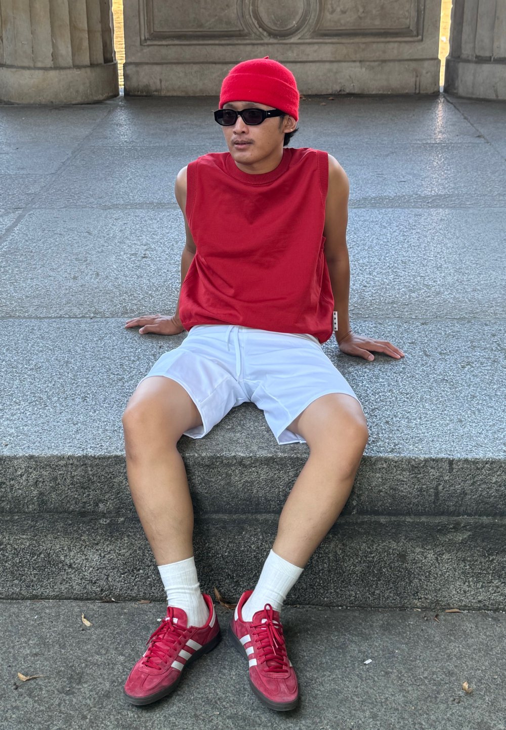 Κόκκινο αμάνικο πουκάμισο, λευκά σορτς, κόκκινα αθλητικά παπούτσια με άσπρες ρίγες και άσπρες κάλτσες. Καθισμένος σε μια πέτρινη σκάλα, με υφή στην επιφάνεια.