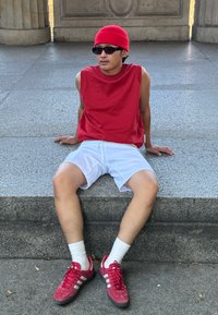 Κόκκινο αμάνικο πουκάμισο, λευκά σορτς, κόκκινα αθλητικά παπούτσια με άσπρες ρίγες και άσπρες κάλτσες. Καθισμένος σε μια πέτρινη σκάλα, με υφή στην επιφάνεια.