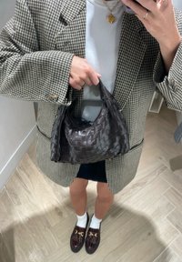 Bolsa de mão de couro trançado castanho escuro com topo arredondado e alça curta, combinada com uma saia preta e um blazer xadrez.