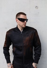 Μαύρο bomber jacket με λαμπερή υφή, διακοσμημένο με διακριτικές καφέ λεπτομέρειες, ριμπ μανσέτες και φερμουάρ μπροστά. Φορεμένο με σκούρα γυαλιά ηλίου.