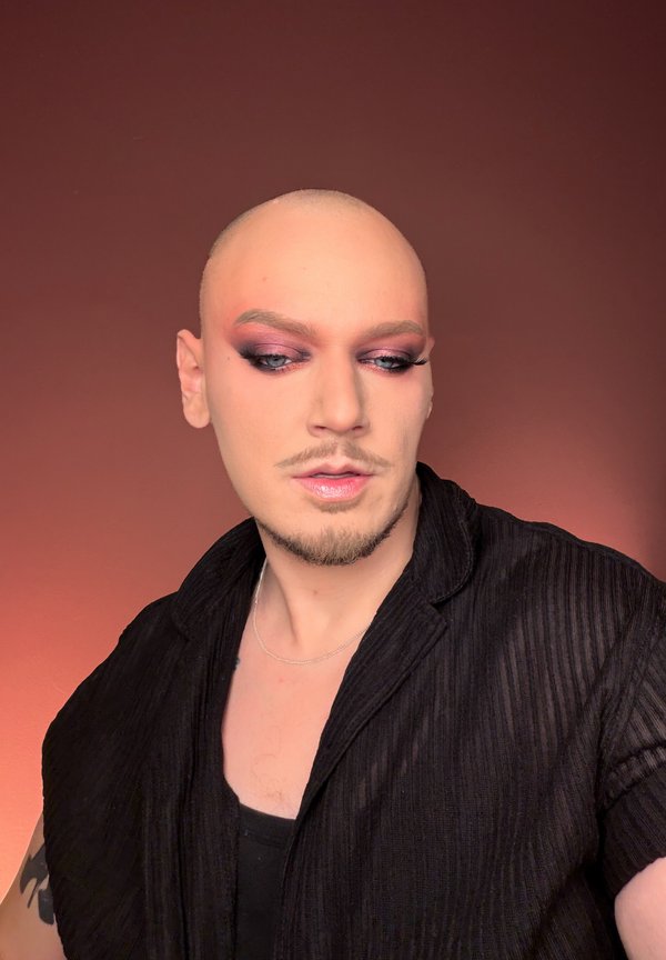 Skaldet person med dristig pink og lilla øjenskygge og blanke læber iført sort skjorte, kigger ned og til siden mod en brun baggrund.