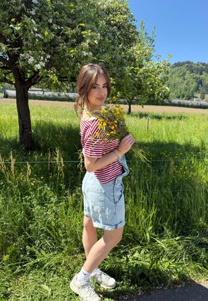 Joven mujer con camiseta de rayas rojas y shorts vaqueros sosteniendo flores silvestres, de pie cerca de un árbol en un campo verde soleado con colinas al fondo.