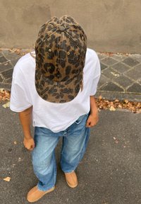 Cappello con stampa leopardata e visiera curva, materiale in cotone, colori marrone chiaro e nero, abbinato a una maglietta bianca e jeans a gamba larga blu.