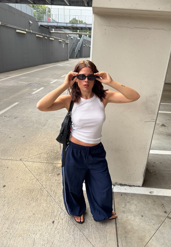 Donna con capelli castani fino alle spalle che si sistema degli occhiali da sole neri, indossa una canotta bianca, pantaloni larghi blu navy, sandali neri e una borsa nera a tracolla.