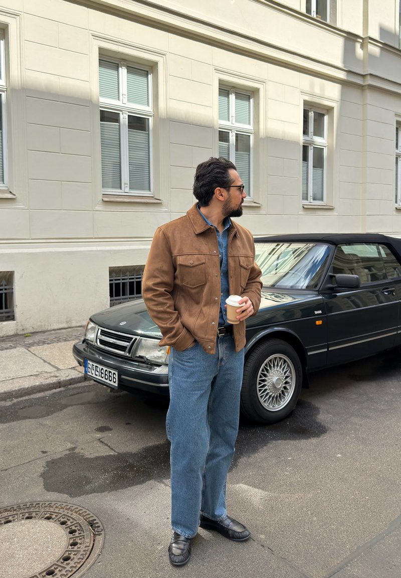 Homme en veste marron et jean bleu tenant une tasse de café, debout à côté d'une voiture décapotable noire dans une rue de la ville.