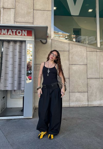 Una joven que lleva una camiseta sin mangas negra, pantalones amplios negros, zapatillas amarillas y collares de cruz se encuentra al lado de una cabina de fotos en una acera urbana.