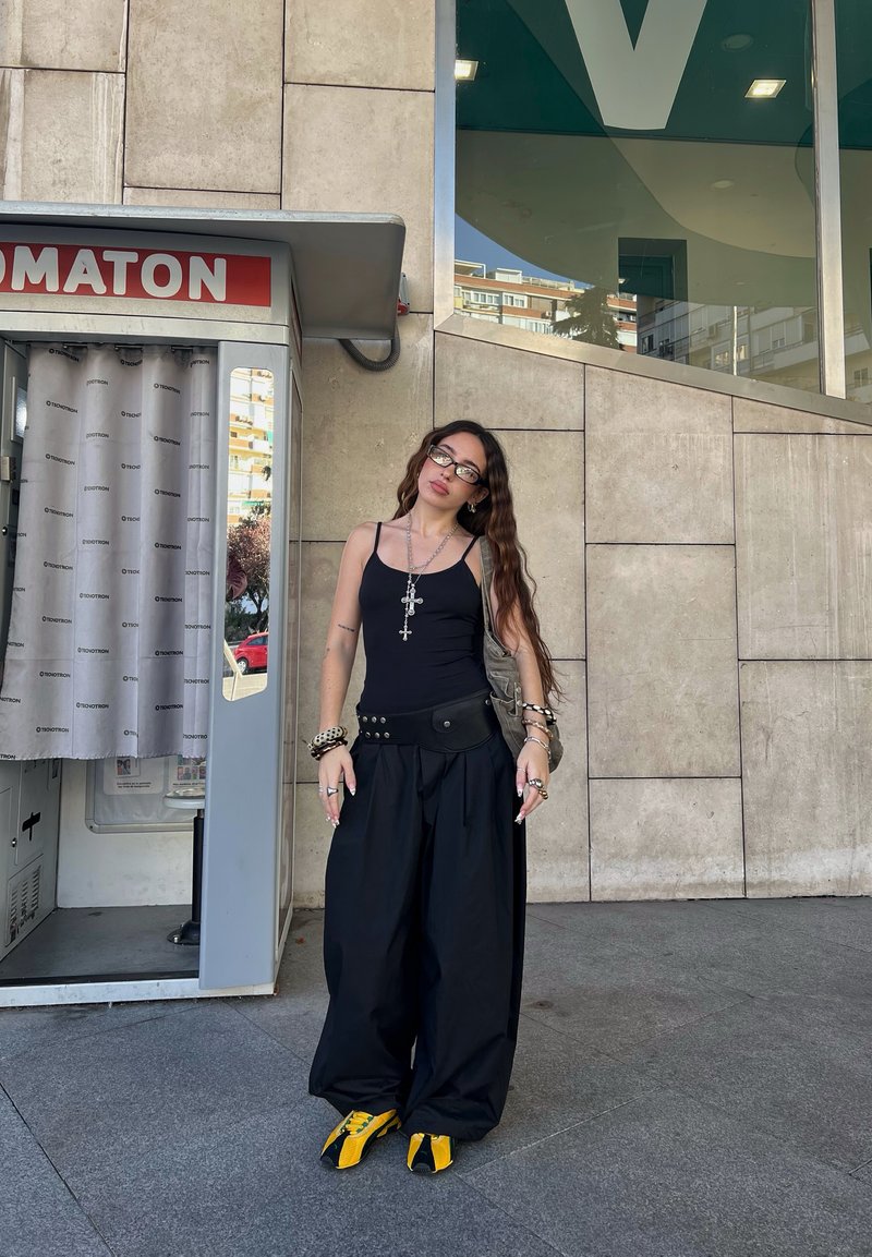 Jovem mulher vestindo uma t-shirt preta, calças largas pretas, sapatilhas amarelas e colares cruzados, ao lado de uma cabine fotográfica numa calçada urbana.